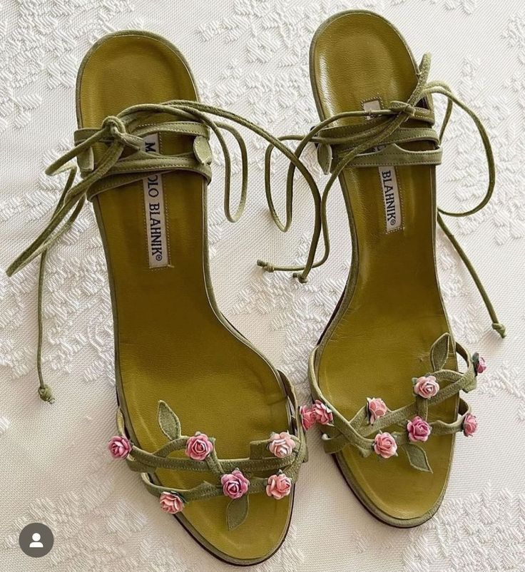 a pair of green manolo blahnik sandals with pink roses and laceup straps are displayed on a white patterned surface. the shoes feature delicate floral detailing and a luxurious design.
