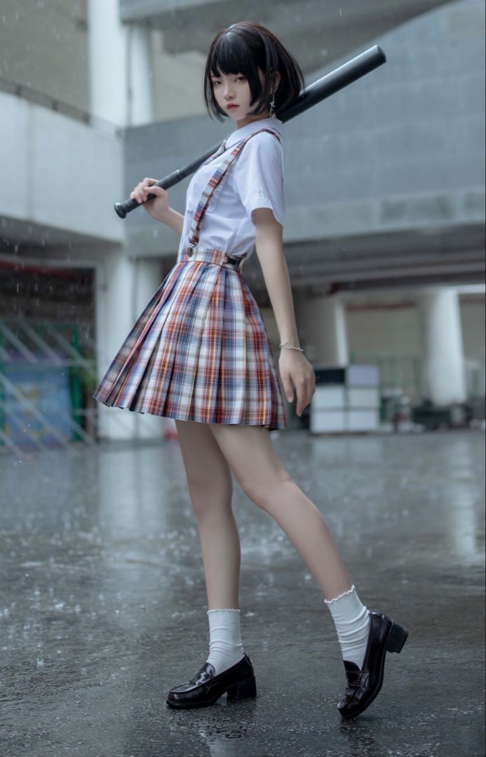 a young woman wearing a japanese school uniform stands on a concrete street, holding a baseball bat in her left hand. she is wearing a plaid skirt, a white shirt and white socks with black shoes. the street appears to be wet, possibly from rain.