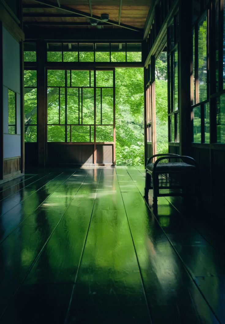 the image showcases a serene and tranquil interior scene within a traditional japanese house.  the space is a long, covered veranda or corridor with large sliding wooden doors and shoji screens that open to a lush green garden. sunlight filters through the foliage, casting shadows and creating bright green reflections on the highly polished dark wooden floor. a simple wooden bench sits quietly near the center of the corridor. the overall atmosphere is peaceful and minimalist, typical of japanese architectural style emphasizing natural elements and simplicity.