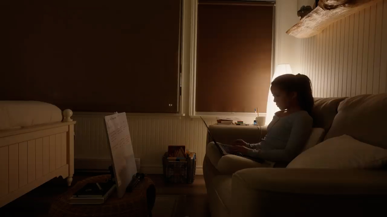 a teenage girl is sitting on a couch in a dimly lit bedroom, illuminated by a nearby lamp.  she is focused on a laptop computer resting on her lap, suggesting she's studying or doing homework in the evening.  the room is cozy with a bed in the background and personal belongings scattered around, adding to the relatable scene of a student's life.
