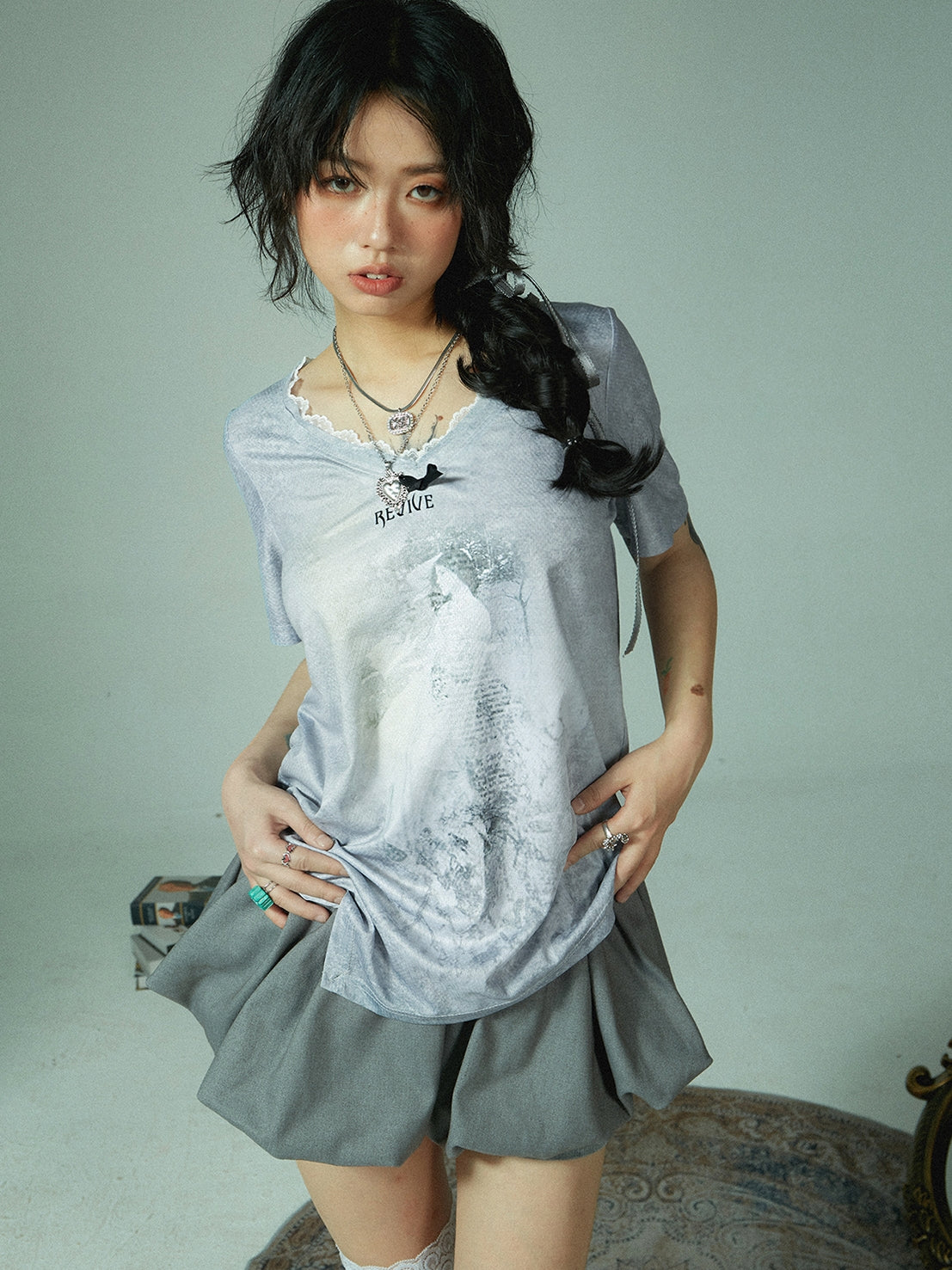 a young asian woman with dark hair styled in a braid is posing in a studio shot. she is wearing a grey vneck tshirt with a graphic print and a matching grey short pleated skirt. she is accessorized with several necklaces and rings. the background is light and neutral, focusing the attention on the model and her outfit. the overall style is casual yet fashionable, with a touch of edgy coolness.  she looks directly at the camera with a slight smile.
