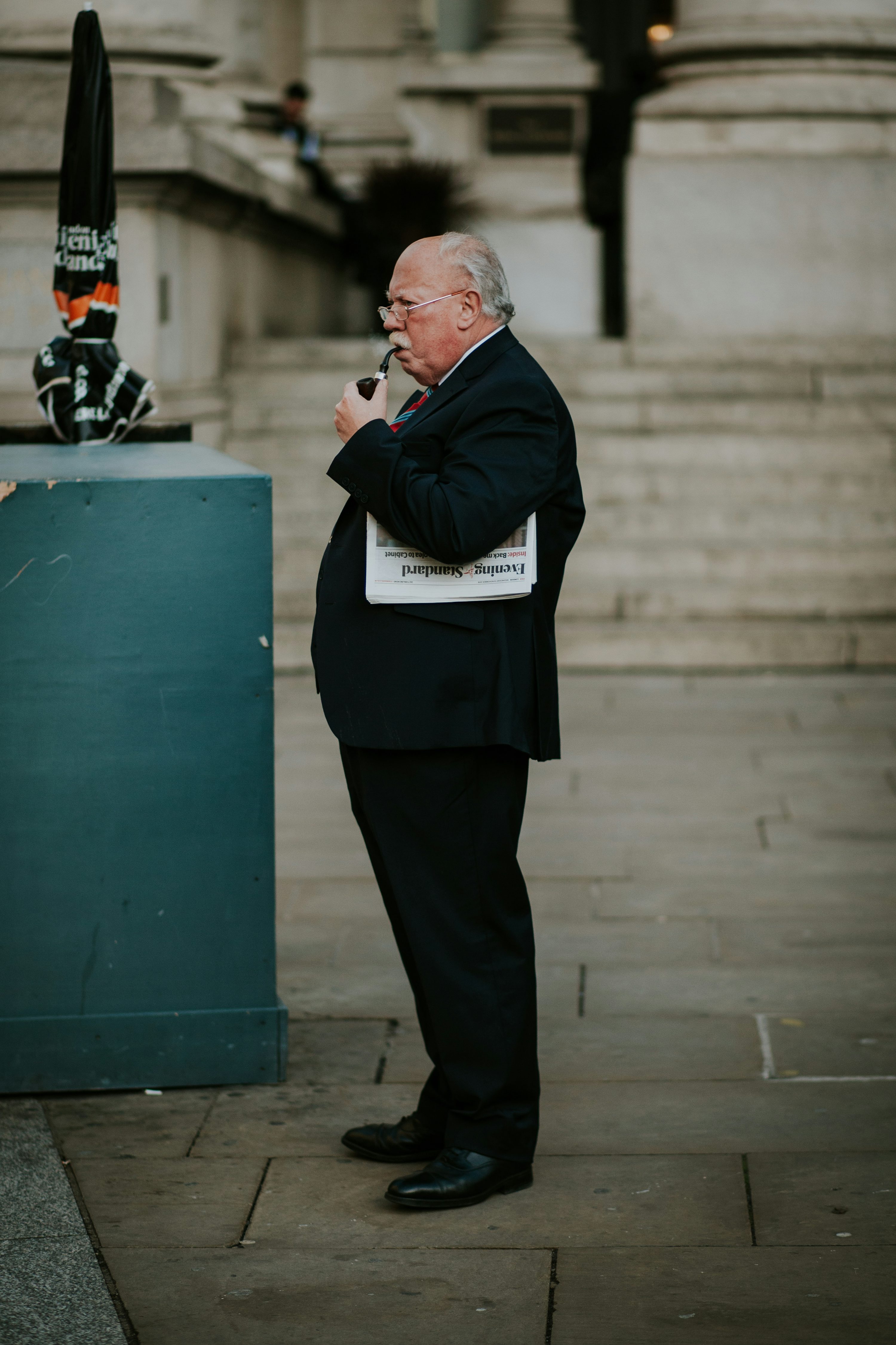 the image shows an older gentleman in a dark suit standing on a city street. he's holding a financial times newspaper under his arm and is smoking a pipe. he appears pensive or thoughtful, with a serious expression on his face.  the background is a blurred image of a stone building facade, suggesting an urban setting. the overall mood is somewhat somber and reflective. the lighting suggests an overcast day.
