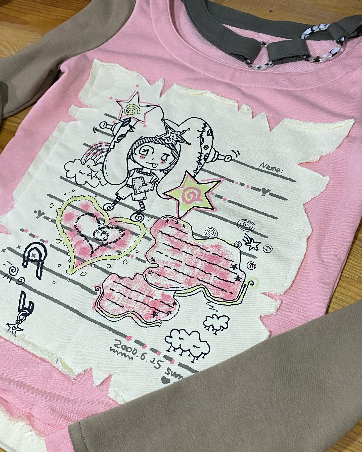 loseup view of a pink longsleeved shirt for a toddler girl. the shirt features a unique, handdrawn style graphic design in a distressed aesthetic. the graphic includes a cute cartoon character, hearts, stars, clouds and other playful doodles. the shirt has a beige/grey raglan sleeve detail.  a name tag area is also visible on the design. the overall style is whimsical and kawaii. the date "2000.6.15 summer" is printed on the shirt.