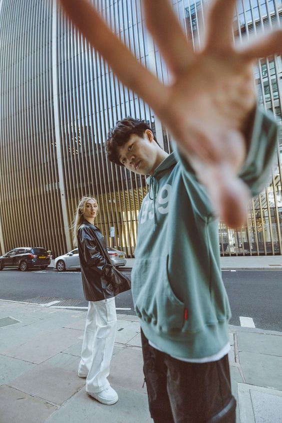 a young man and woman stand on a city sidewalk, a towering skyscraper rising behind them.  the man, wearing a green hoodie and black pants, stares directly at the camera. his hand is outstretched, partially obscuring the lens. the woman, in a black leather jacket, white pants, and white sneakers, gazes off to the side. cars are parked and moving along the street behind them. the photo is taken in daylight, giving the image a natural and candid feel.