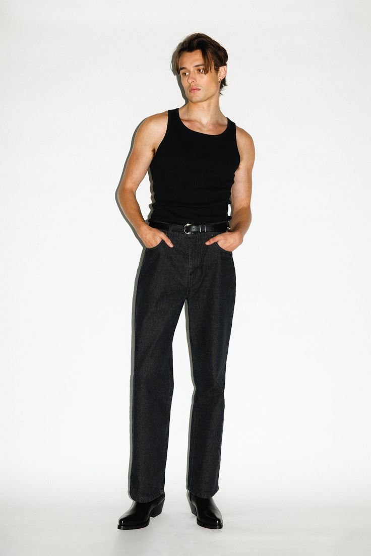 a fullbody shot of a man standing against a white background. he's wearing a black tank top, darkwash denim jeans, a black belt, and black cowboy boots. his hairstyle is short and slightly tousled.  the image is a fashion shot, showcasing the clothing. the man looks directly at the camera with a serious expression.
