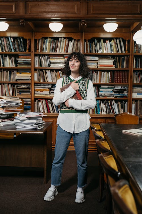 a woman stands in a library, smiling while holding a book in her arms. she's wearing a patterned vest over a white shirt, jeans, and white platform shoes. behind her is a large bookshelf filled with books. there is a table on either side of her, one with a stack of papers and the other next to some chairs.