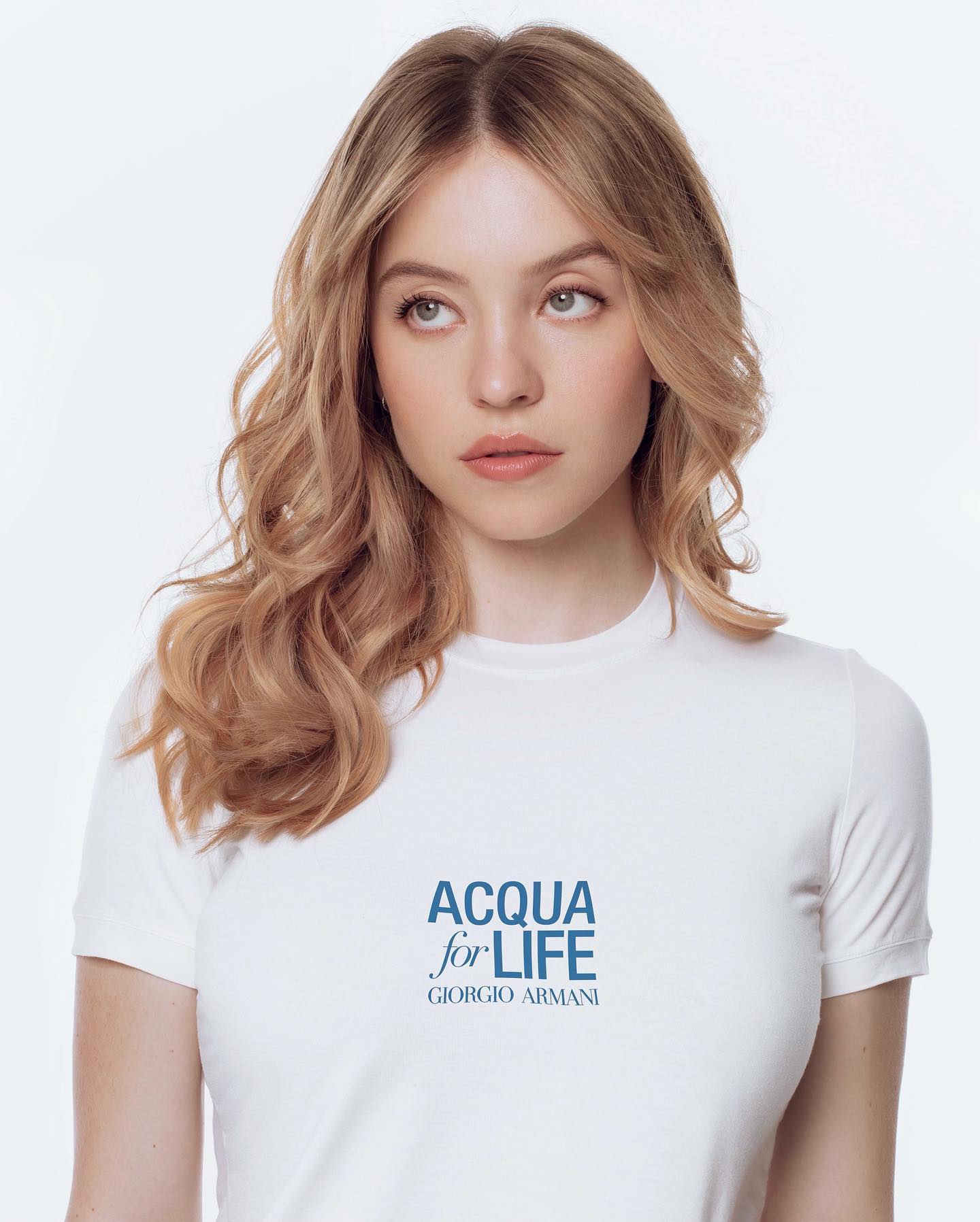 closeup portrait of a young blonde woman with wavy hair, wearing a white tshirt with the "acqua for life giorgio armani" logo. she is looking off to the side with a thoughtful expression. the image is a studio shot against a plain white background, suggesting a fashion or advertising campaign for the charity.