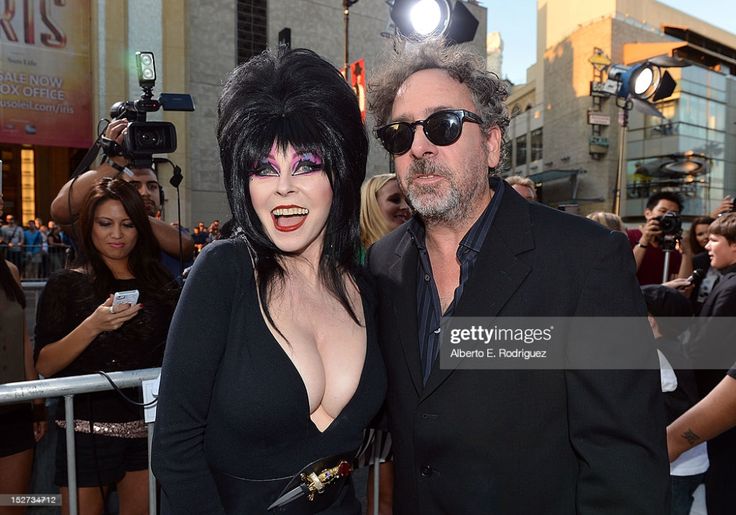 a photo of actress carolyn jones and director tim burton on the red carpet at a film premiere. jones is wearing a black dress with a plunging neckline and her signature black hair and makeup. burton is wearing a dark suit. they both have big smiles on their faces and are surrounded by photographers.