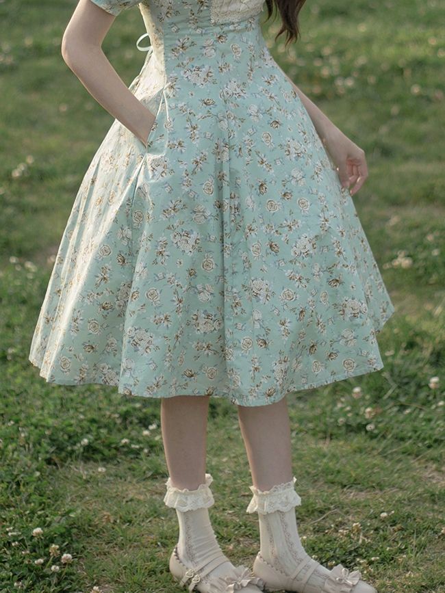 a woman in a vintageinspired, light blue floral dress stands in a field. the dress is midi length with a full skirt and short sleeves. she's wearing white socks and shoes. the image is taken at eye level and bathed in soft, natural light, giving it a dreamy and romantic feel.
