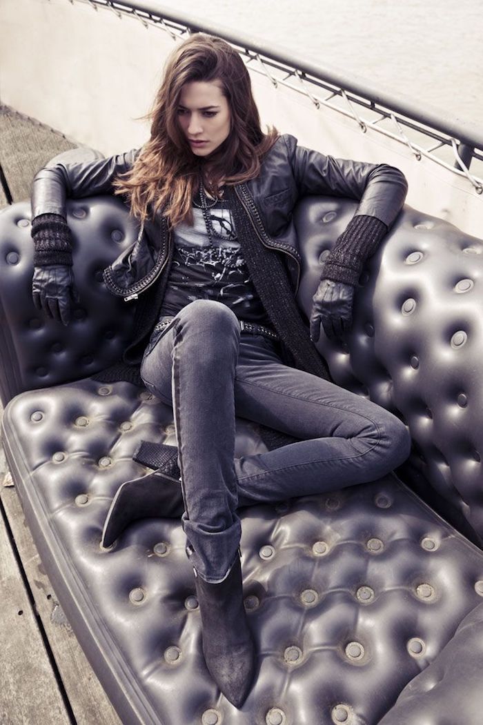 a young woman with long brown hair is sitting on a black leather couch outdoors. she is wearing a black leather jacket, dark jeans, and black ankle boots. she is accessorized with gloves and a necklace. the couch is tufted and appears to be made of a leatherlike material. the overall aesthetic is stylish and chic.