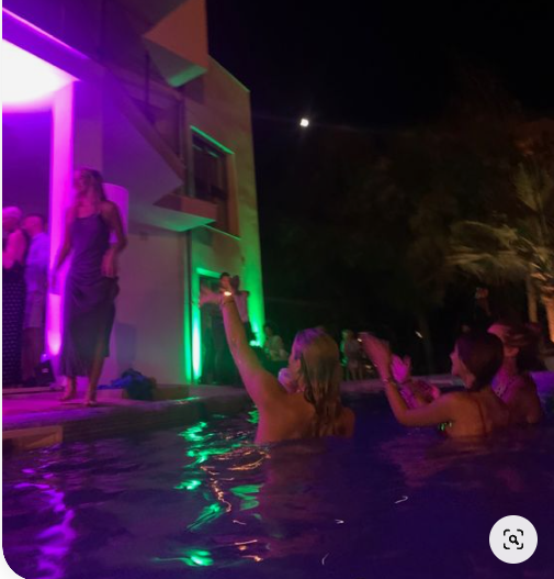 a group of women are having a party at night. they are swimming in a pool and there is a house in the background. the house and pool are lit up with colorful neon lights. the overall atmosphere is festive and fun.