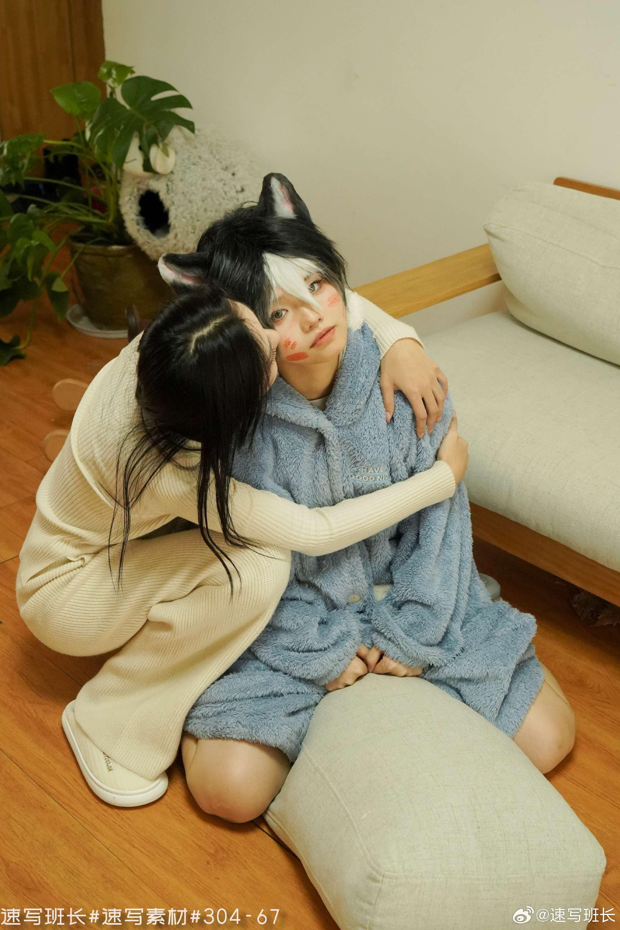 two women are in a living room. one woman is sitting on a pillow wearing a blue bathrobe and cat ears. the other woman is hugging her and kissing her on the cheek. there is a plant and a plush toy in the background.