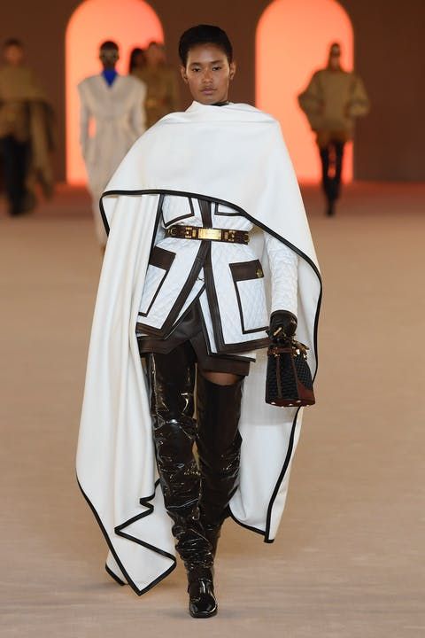 a female model walks the runway in a white cape coat, black overtheknee boots, a black and white quilted jacket, a brown leather belt, and a small black handbag.  the model is walking on a white runway and the background is a white and orange wall.
