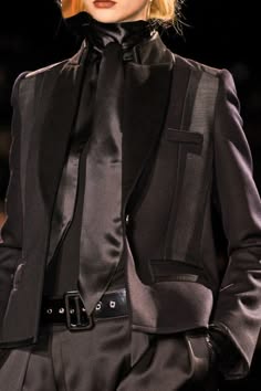 a woman in a black blazer with a black tie and black leather belt posing in a studio setting for a photoshoot.