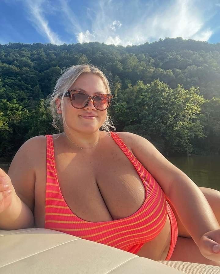 a woman with blonde hair and sunglasses is lounging on a boat wearing a pink and orange striped swimsuit. behind her are a lake, trees, a mountain, sky, and clouds.