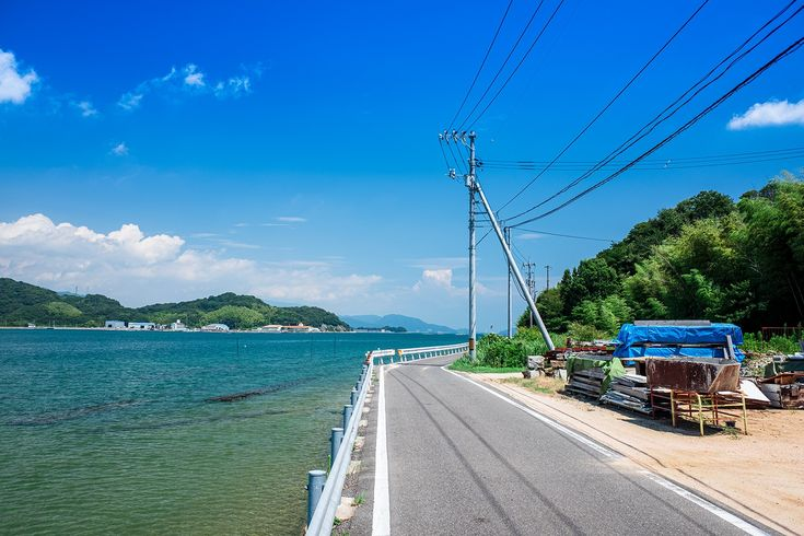a scenic view of a road running alongside the ocean on a clear day. the sky is blue with scattered clouds. on the right side of the road are trees, a utility pole with power lines, and some debris covered by a blue tarp. in the distance, there's an island. a guardrail separates the road from the ocean on the left.