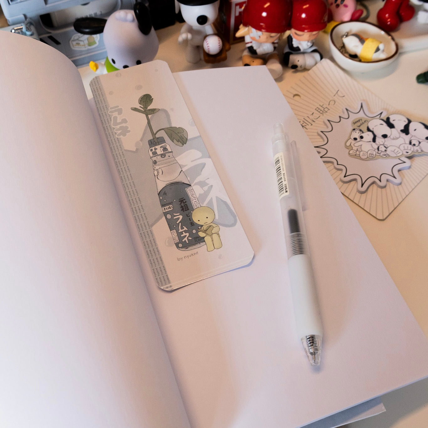 a white notebook lays open on a table with a pen, bookmark, and other stationary items. the bookmark has a cute cartoon illustration of a soda bottle with a plant growing out of it. the bottle says "ラムネ" which is the japanese word for soda.  the bottle is grey with blue text and the character is light grey. there is a white background behind the bookmark. the notebook is white and has a smooth surface.  the pen is white with a dark grey ink tip.