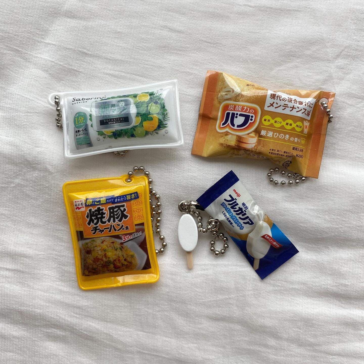 the image shows four different packaged food items attached to keychains on a white surface. there is a sabrino botanica face mask package, a papa package, a charhan fried rice package, and a meiji bulgaria yogurt package.