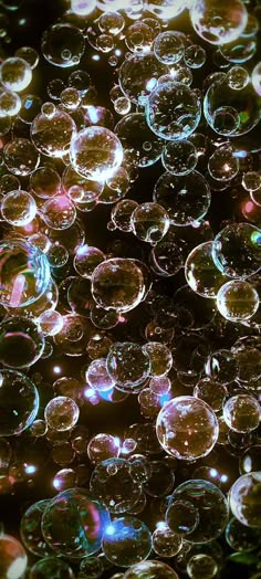 a closeup shot of a large cluster of iridescent bubbles, mostly out of focus, set against a dark background. the bubbles are of various sizes and shapes, and they reflect light in a variety of colors. the overall effect is abstract and dreamy, with a sense of magic and whimsy.