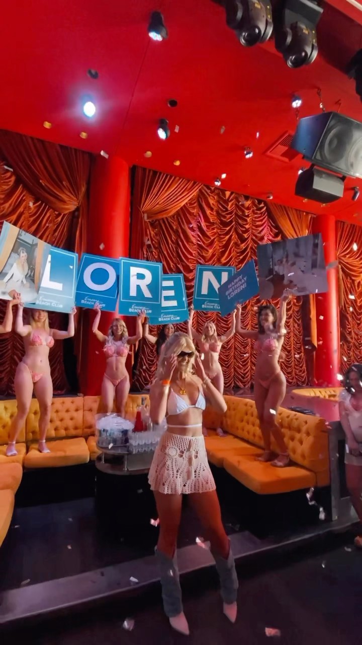 a group of women are celebrating, possibly at an event or party inside what looks like a nightclub. some women are wearing bikinis and holding signs that spell out "loren beach club". one woman stands in front wearing a white bikini top, a crocheted skirt, and boots. confetti is falling.