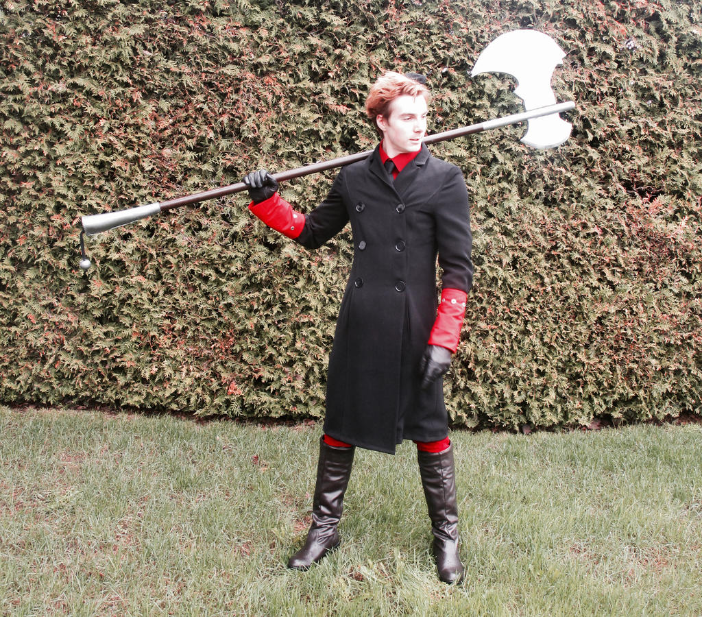 a man in cosplay is standing on a lawn with a hedge behind him. he is wearing a long black coat, a red shirt, red gloves with black gloves over them, and black boots. he is holding a large, silvercolored axe over his shoulder.