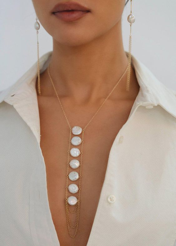 a closeup shot of a woman wearing a delicate gold necklace with dangling freshwater pearls and matching earrings. she is dressed in a crisp white buttondown shirt, accentuating the elegance and simplicity of the jewelry. the soft lighting enhances the natural beauty of the pearls and the model's skin.
