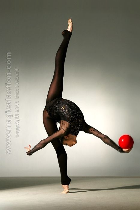 a female gymnast is balancing on one leg with her other leg extended behind her. she is holding a red ball in her right hand. she is wearing a black leotard and tights. the background is a plain gray. she is practicing a gymnastic routine.