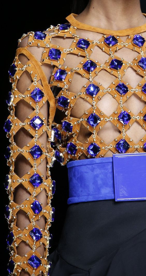loseup shot of a woman's torso wearing a stunning haute couture dress. the dress is a rich, earthy brown suede with a cutout design intricately embellished with numerous dark blue gemstones and gold accents.  the gemstones are set in a gridlike pattern, creating a striking contrast against the suede.  a vibrant blue leather belt cinches the waist. the overall style is luxurious and glamorous. the image highlights the meticulous detail and craftsmanship of the garment.