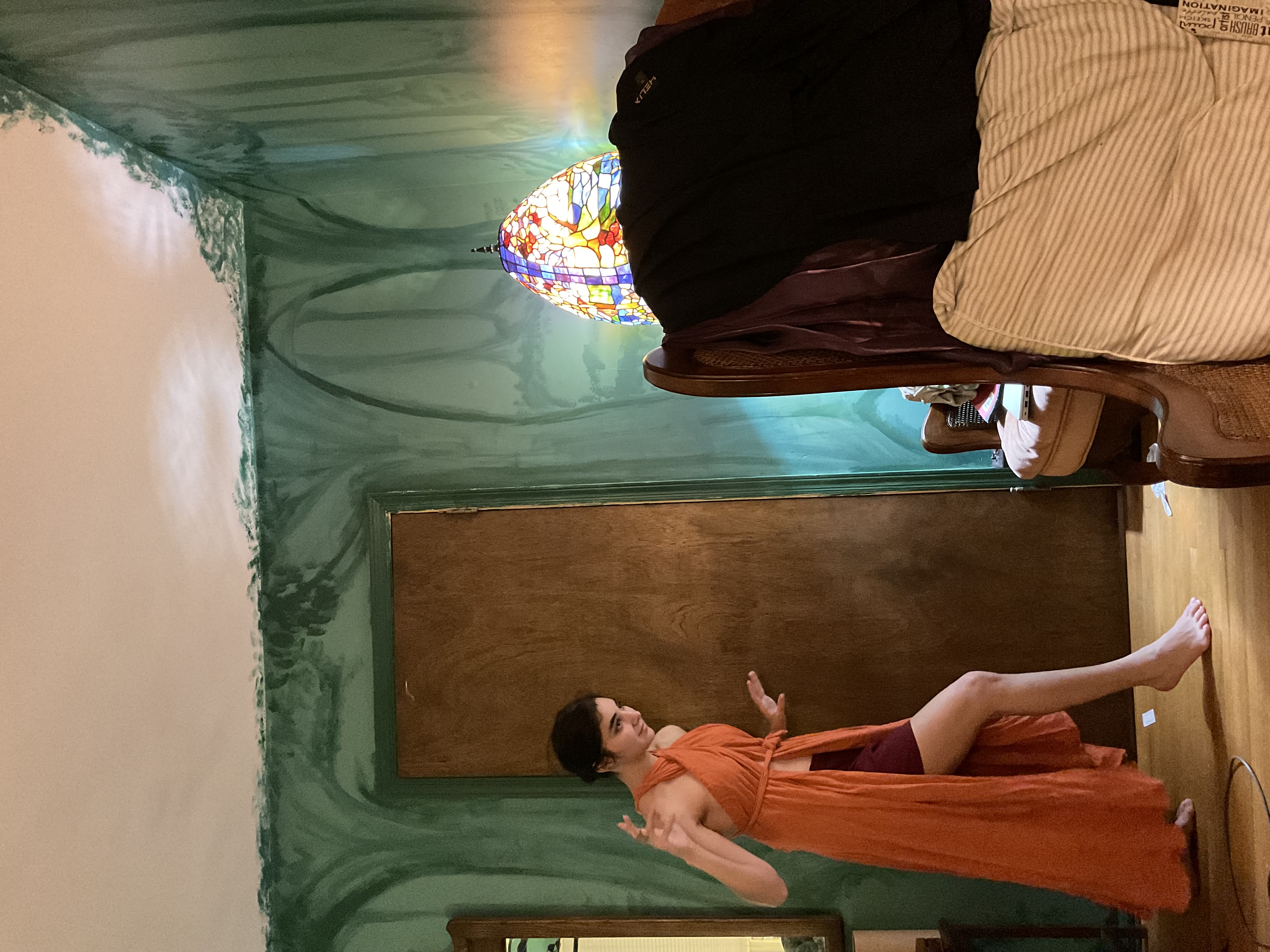 a young woman with dark hair is standing in a room with a uniquely painted green wall depicting a forest scene. she is wearing a flowing orange wrap dress and is posing with one arm raised.  a stainedglass lamp sits on a wooden chair in the background, and the overall ambiance of the room is cozy and artistic. the woman is barefoot and the floor appears to be hardwood. the image appears to be a candid portrait of the woman in her home.