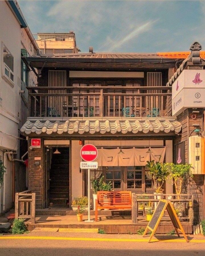 the image shows the facade of a building, possibly a cafe or coffee shop, in an urban setting. the architecture has asian, possibly japanese or korean, influences. there's a bench in front of the building, an awning above the entrance, and signage indicating a cafe. the scene appears to be a street view in a town.