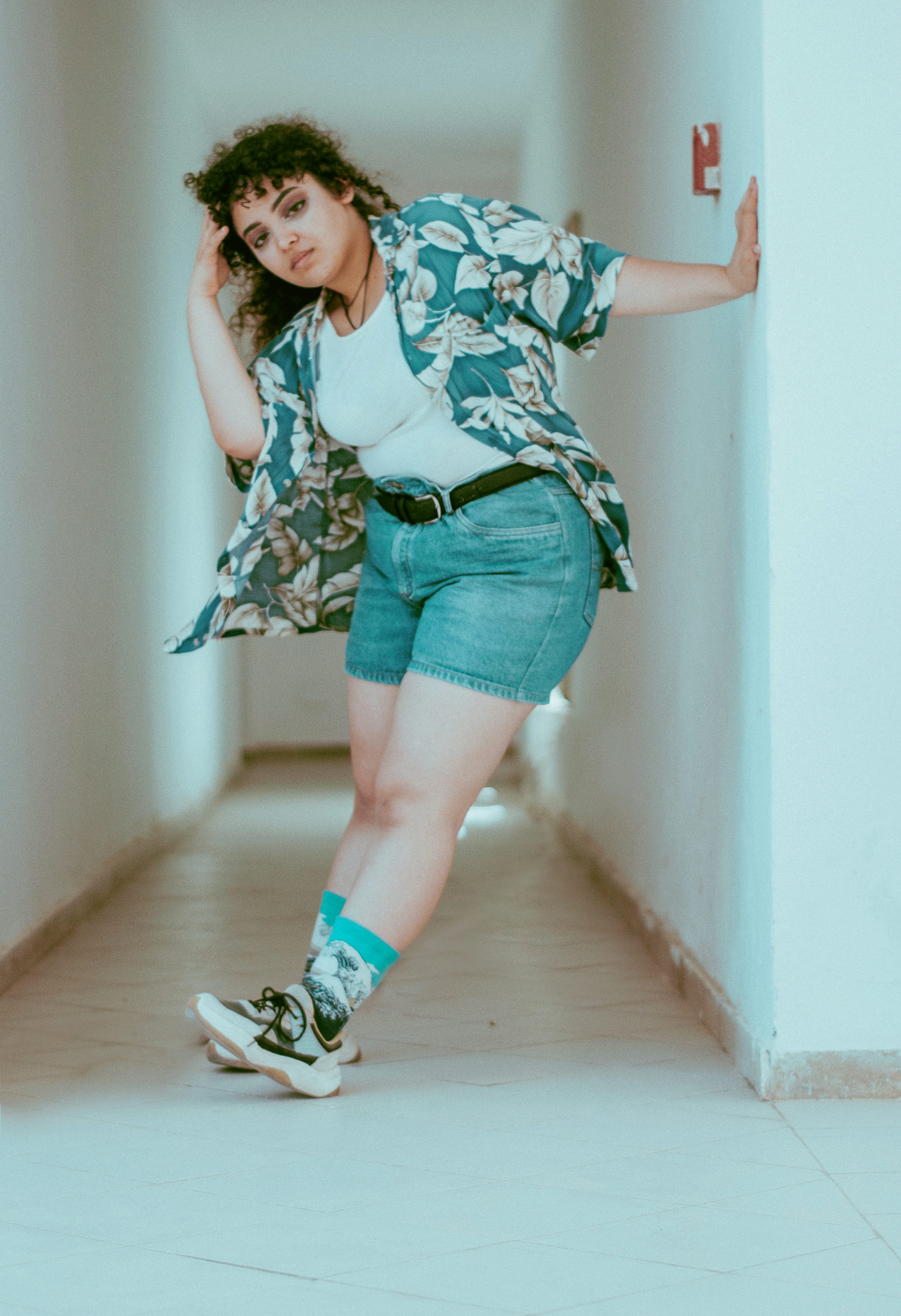a young woman with curly hair is posing in a hallway. she is wearing a teal floral shirt, denim shorts, white socks with a design, and white sneakers. she is leaning against a wall and looking at the camera. the hallway is white and bright.