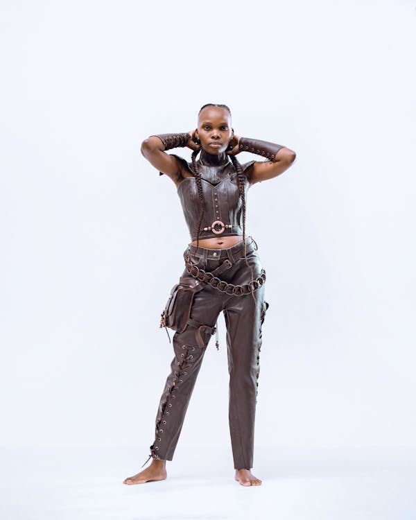 a woman stands barefoot in a studio, posing with her hands behind her head. she is wearing a brown leather outfit consisting of a sleeveless top with a high collar, arm warmers, and pants. the pants have a laceup detail on one leg and a pocket attached to the side. a chain belt adorns her waist. her hair is styled in braids. the background is a plain white.