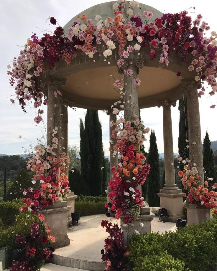 a gazebo is heavily decorated with a vibrant floral arrangement featuring pink, red, and purple flowers, including roses. the gazebo has stone pillars and is situated outdoors in a garden setting, with trees and bushes in the background. the scene appears to be set for a wedding or ceremony.
