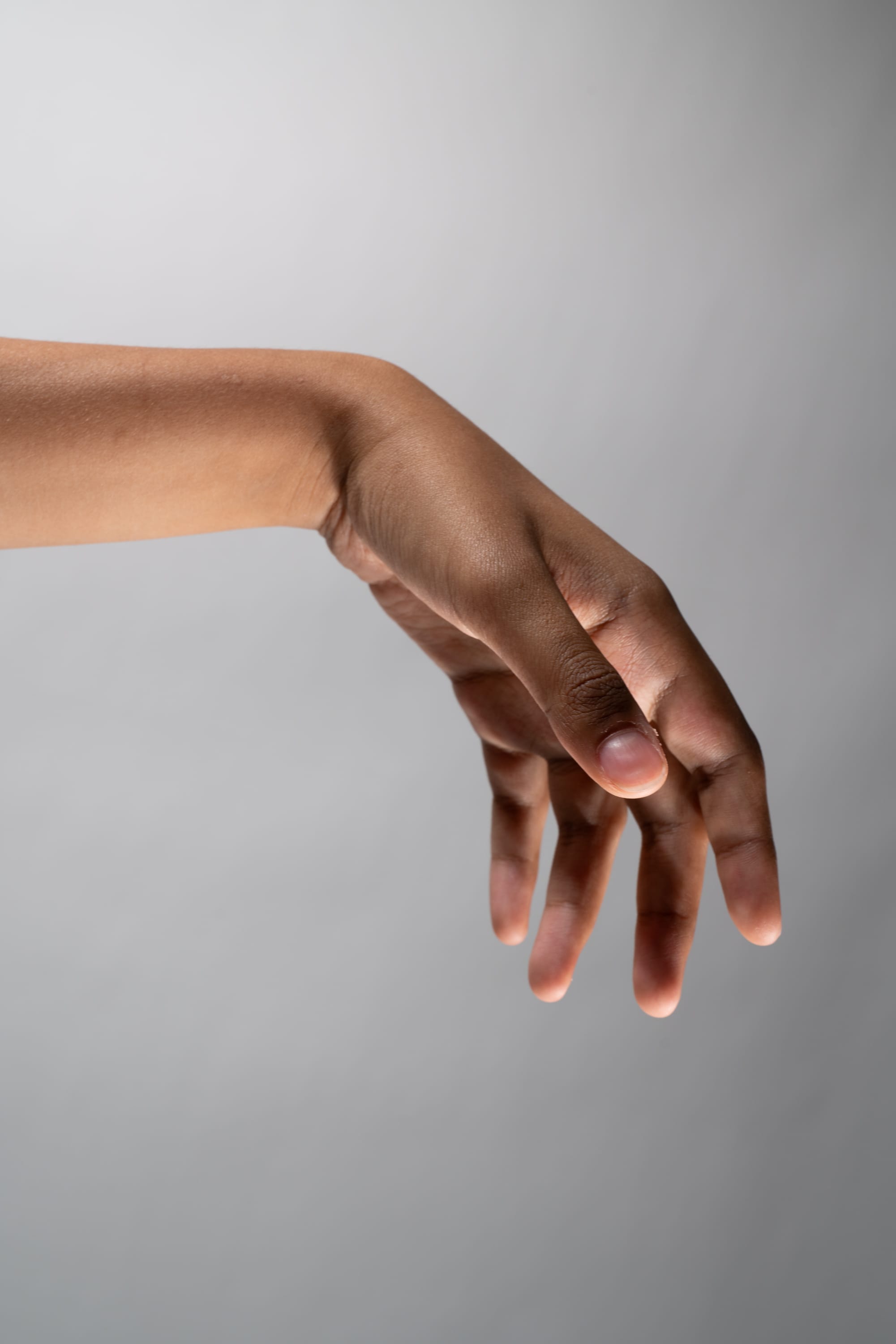 the image showcases a single hand, belonging to a person with dark skin. the hand is reaching downwards, with the fingers slightly curled inwards.  the backdrop is a neutral gray, which emphasizes the contrast of the hand's color and texture.  the lighting is soft and diffused, creating a subtle shadow beneath the hand.