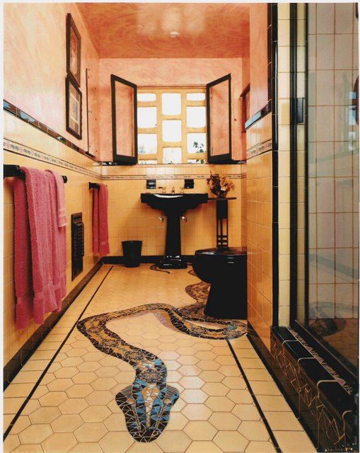 the image shows a bathroom with pink and yellow tiled walls and a hexagonal tiled floor. there is a black toilet and sink. the window is open and a snake mosaic is on the floor. there are pink towels hanging on a rack.
