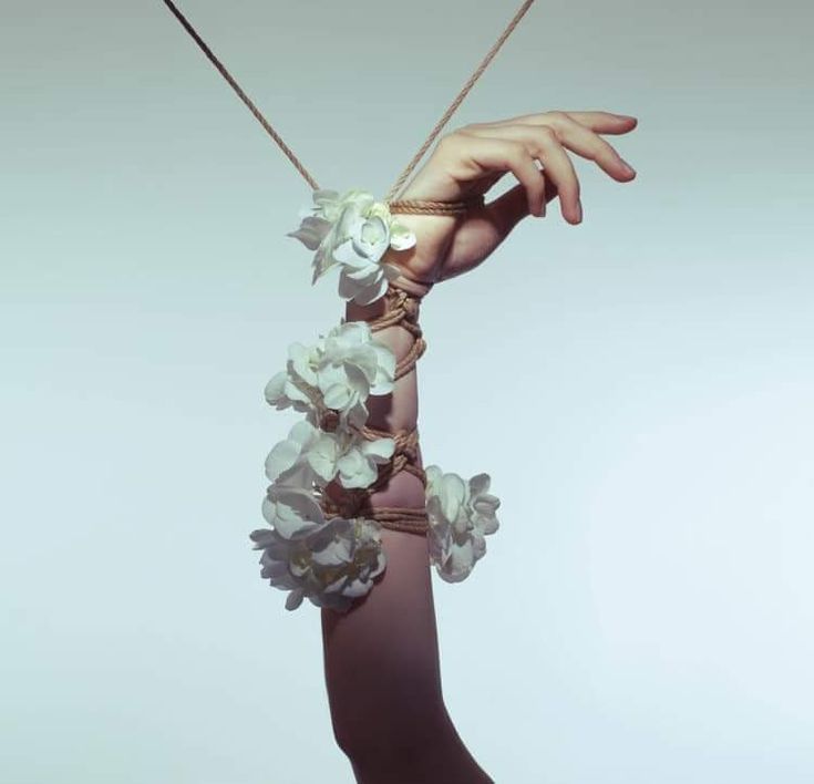 the image is a stylized photograph of a woman's arm and hand bound with rope. the rope is intricately woven around the arm, and bunches of small, white flowers are incorporated into the bindings. the overall effect is elegant and surreal,  creating a contrast between the delicate flowers and the restrictive nature of the rope. the lighting and color palette contribute to a soft, ethereal atmosphere. the image is likely intended to be conceptual or artistic, exploring themes of restraint, beauty, and perhaps even vulnerability.