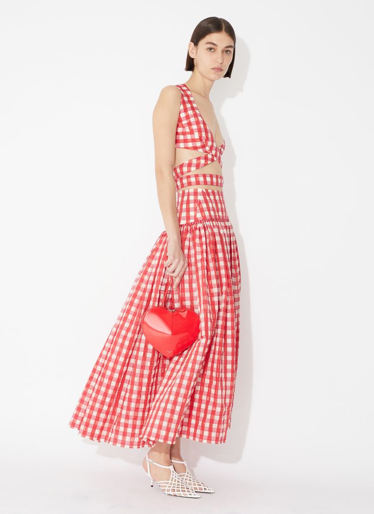 a model poses in a full shot, wearing a red and white checkered dress consisting of a cutout top and a maxi skirt. she holds a red heartshaped purse and wears white caged heels. the background is a white studio backdrop.