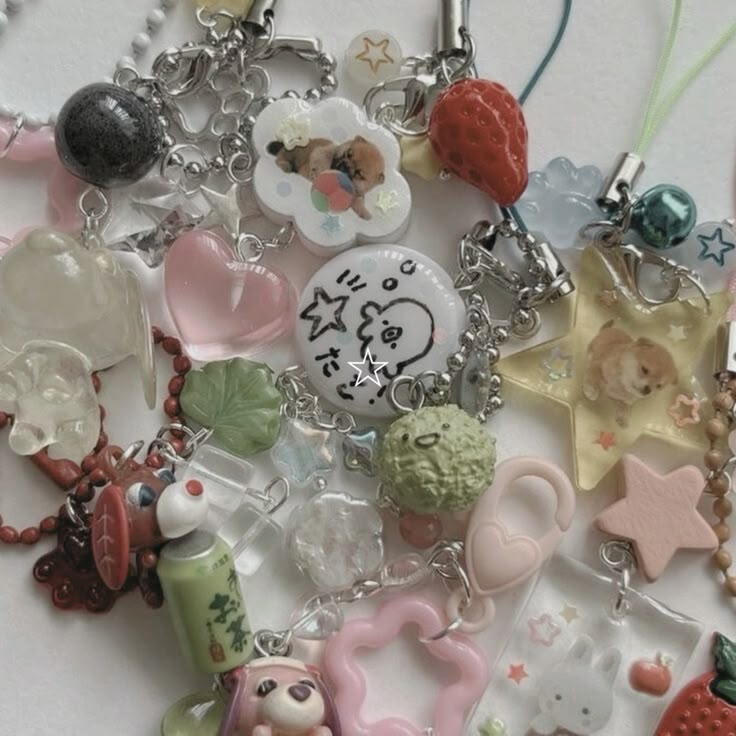 a closeup overhead shot of a collection of various charms and keychains. there are numerous plastic, beaded, and kawaiithemed trinkets arranged in a pile, including charms of cartoon characters, stars, hearts, a strawberry, clouds, dogs, bunnies, a can, a leaf, and textured balls. colors are primarily pink, green, white, red, and clear with silver hardware.