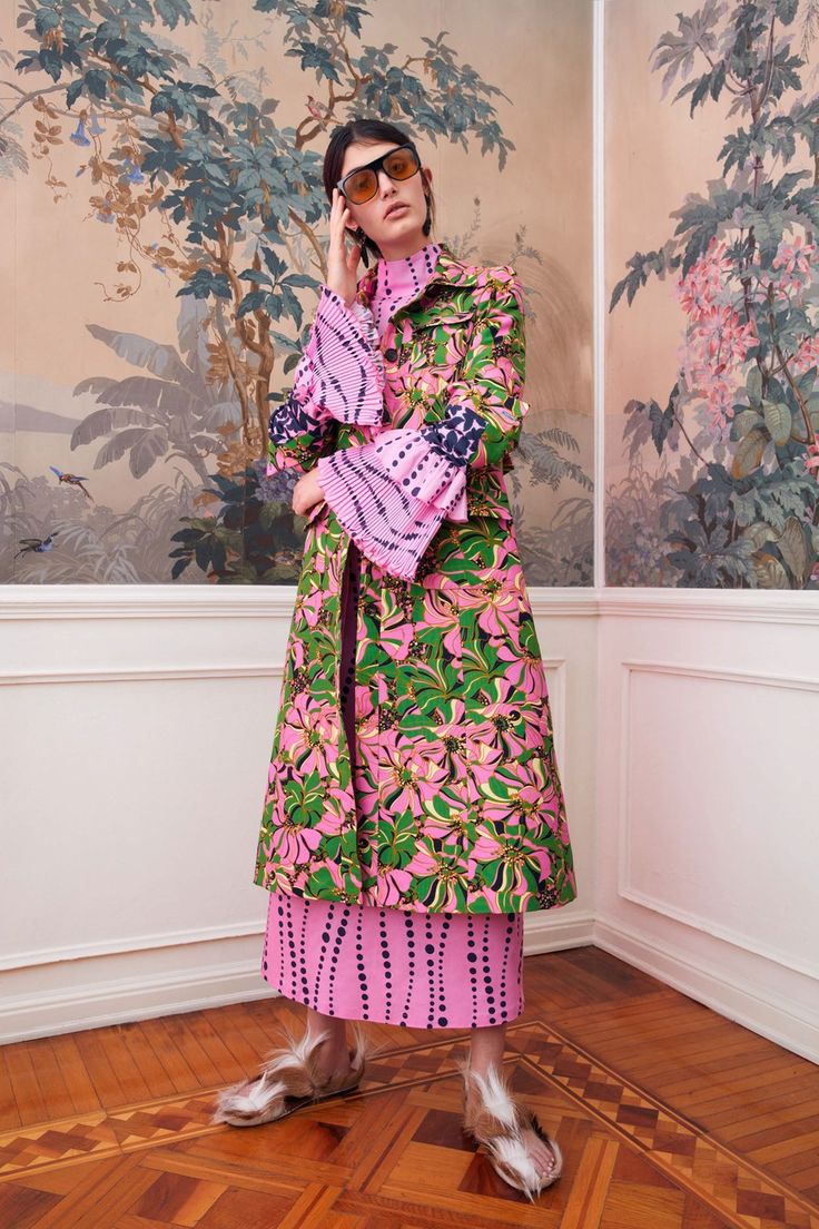 a full shot of a fashion model wearing a pink and green floral coat and matching skirt. the coat has pleated sleeves and the skirt has a dotted pattern.  she is wearing fur slippers and sunglasses. the model is posing in a room with ornate wallpaper and a wood floor. the overall aesthetic is elegant and sophisticated.