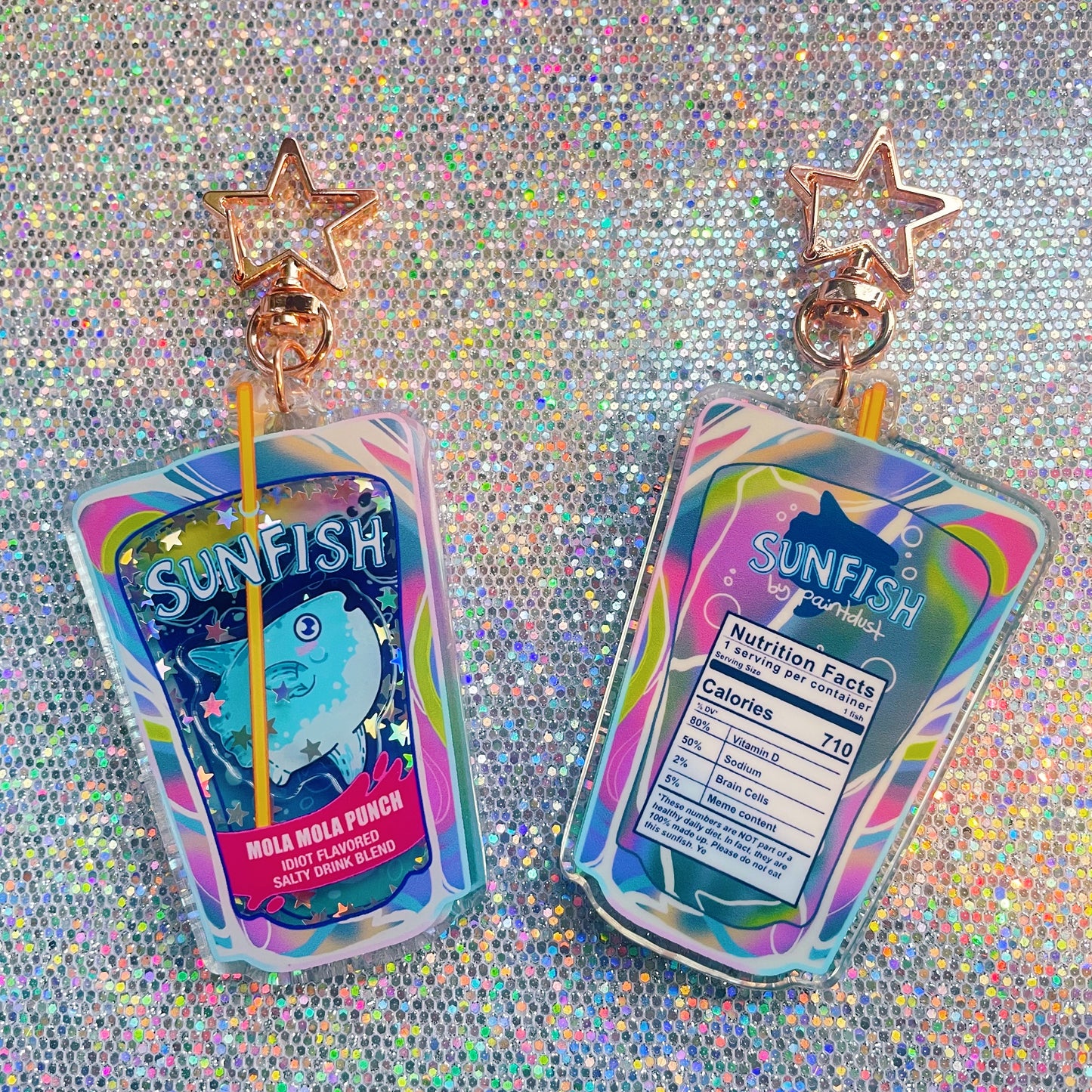 he image shows two resin keychains shaped like juice boxes. each keychain features a design with a cartoon sunfish, a playful "nutrition facts" label with humorous details, and a pastel iridescent color scheme.  the keychains are adorned with glitter and have a starshaped charm attached to a rose gold colored ring.  they are displayed against a sparkly, iridescent background. the overall aesthetic is kawaii and quirky.