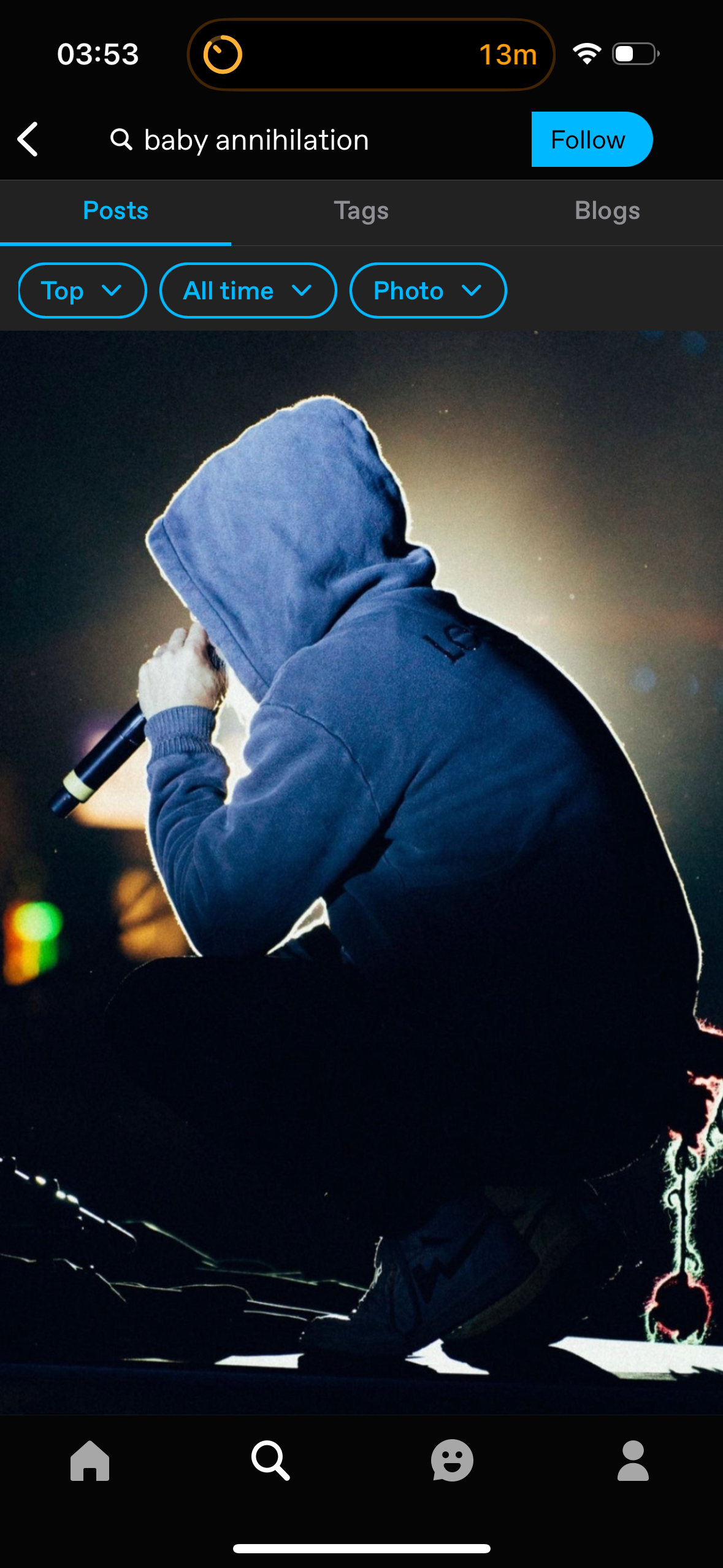 a male rapper, wearing a blue hooded sweatshirt, performs on stage holding a microphone. the image is dark and stylized with only the rapper and the microphone in focus, with the background blurred and dark.  the image has a strong sense of motion and energy, suggesting the excitement of a live performance.