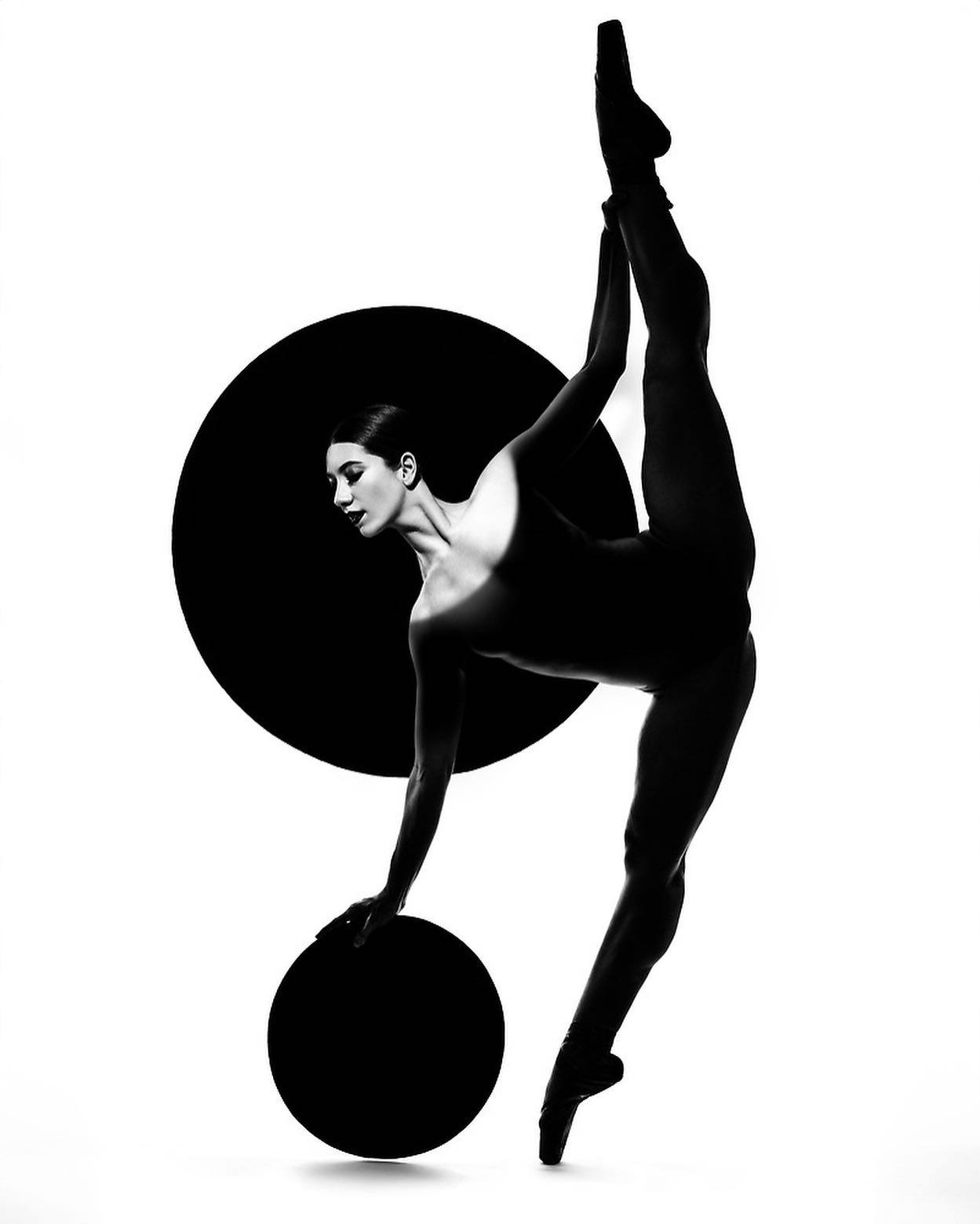 a black and white photo of a ballerina in a contemporary pose. she is positioned with her back to the camera, her right leg is bent backwards and her left leg is straight out in front of her. she is holding onto a black disc with her left hand, and her right hand is stretched out towards the side of the frame. her body is completely silhouetted against a white background, and the black disc is in the background. the ballerina is looking to her left side. the photo is a striking and powerful image of a female athlete.