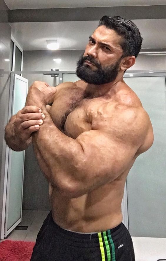 a muscular man with a beard flexes his biceps in a gym locker room, showing off his massive physique. he is shirtless and wearing black athletic shorts with green stripes. the photo is taken from the chest up and captures the intensity and focus of a bodybuilder.