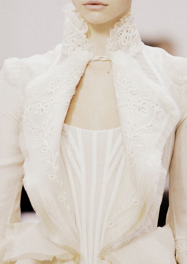 a closeup shot of a woman wearing an intricately designed white dress with lace and embroidery details. the dress features a high collar with delicate ruffles and a corsetstyle bodice with vertical stripes. the overall aesthetic is elegant and reminiscent of a wedding dress or highfashion garment.