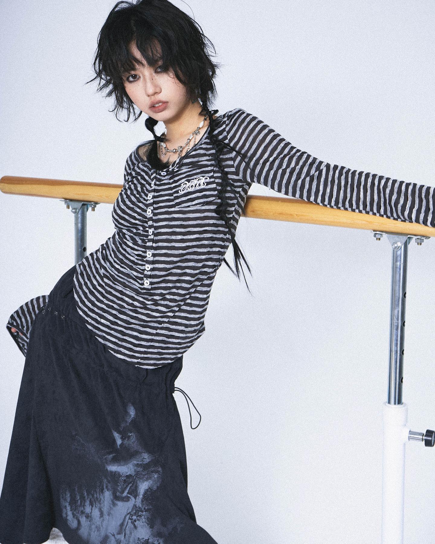 a young woman with short, dark, choppy hair is modeling a dark grey and black horizontally striped longsleeved shirt with small buttons down the center. she's also wearing a long, darkcolored skirt with a subtle print.  she has on a delicate necklace. the woman is posing with her arm resting on a ballet barre in a studio setting against a lightgrey background. her style is edgy and alternative, leaning towards grunge or goth aesthetics.