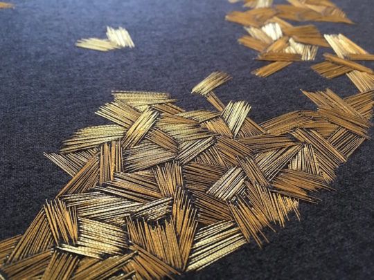 closeup of a dark fabric surface with intricate gold embroidery. the embroidery features an abstract pattern of short, parallel lines, creating a textured effect. the lines are arranged in clustered groups, with some areas more densely embroidered than others. the overall impression is a luxurious and artistic textile design.