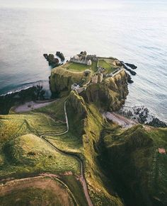 an aerial view of dunnottar castle in scotland, perched on a cliff overlooking the sea. the ruins of the castle are visible on the grassy cliff top, with a winding path leading up to it from the green, hilly landscape. the coastline is rocky, with the blue sea stretching out to the horizon.