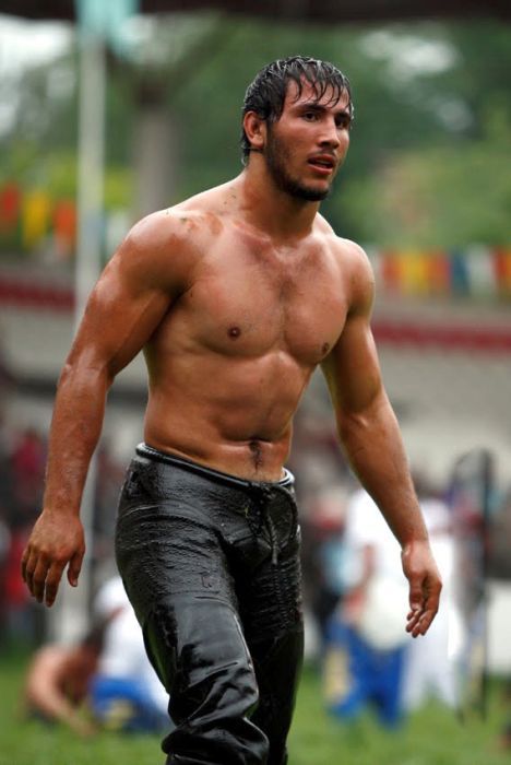 a shirtless muscular man in black oil wrestling pants stands on a green grass field. he has a six pack and oiled skin. he is gripping his hand with his left hand and looks at the camera. this is most likely a wrestler participating in a competition.