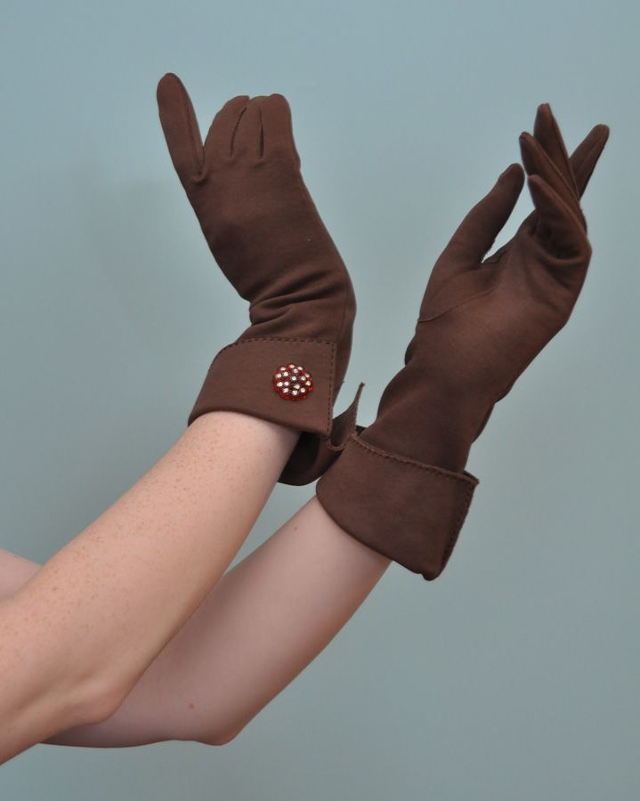 a woman is wearing brown gloves and has her hands up in the air, one hand is closer to the camera than the other. she is wearing brown gloves that have a button on one of the gloves. her hands are in a very feminine pose, with the fingers spread out and the palms facing the camera. the background is a plain blue wall.  the gloves are elegant and stylish.  the model's arms are long and slender.  the image is welllit and the colors are vibrant.
