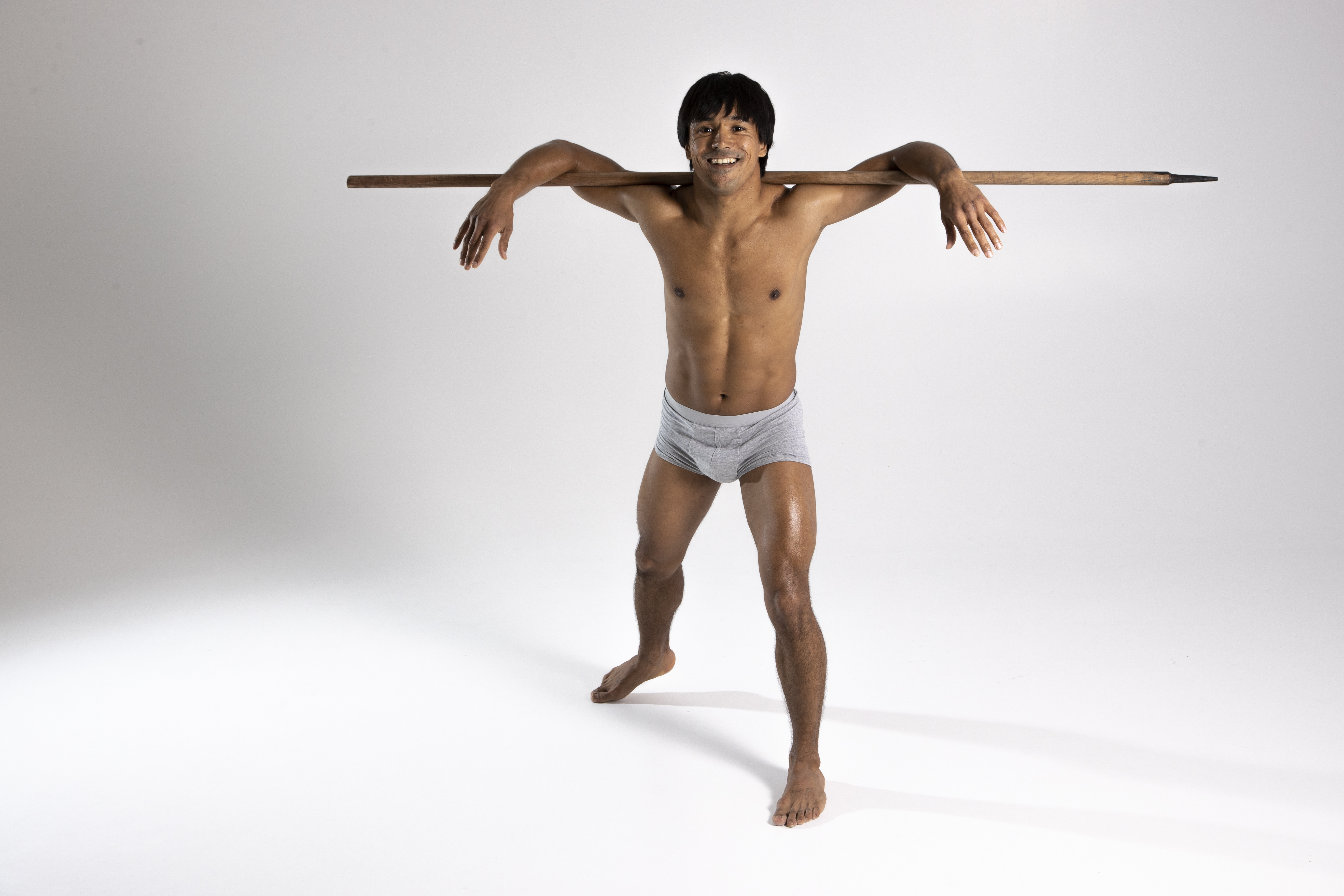 a shirtless man in gray underwear stands barefoot in a studio against a white background. he smiles as he balances a wooden spear across his shoulders. his skin has a sheen as if it is oiled.